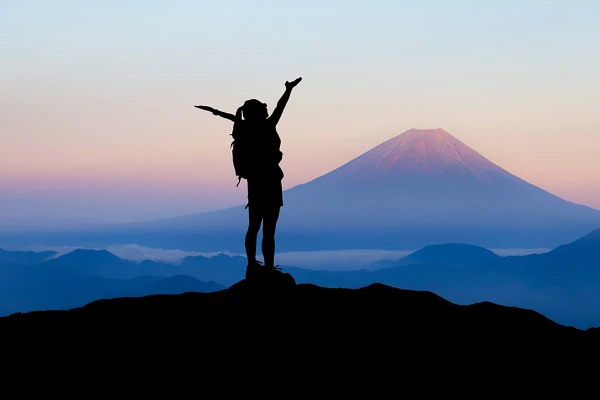 A hiker triumphantly raises their arms atop a mountain with Mount Fuji in the background at dawn.